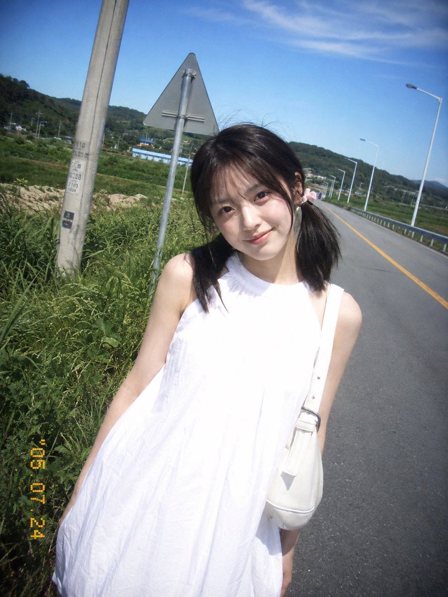 复古 CCD 卡片机夏日旅行抓拍，一位20岁出头的年轻东亚女idol站在乡间公路边。黑棕色低位双马尾，细碎刘海，浅粉色发圈，淡妆，冷白皮，真实…