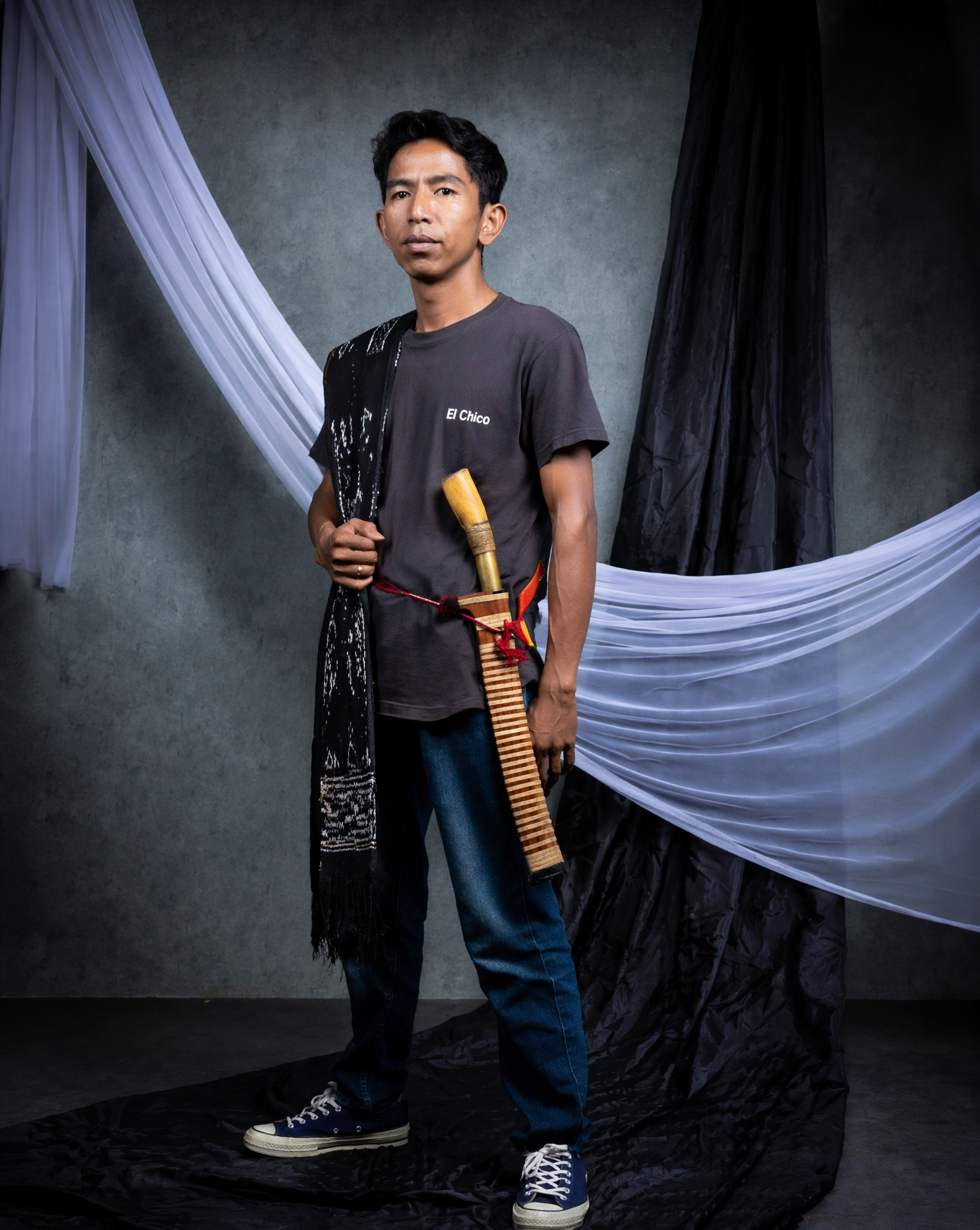 A Southeast Asian man stands centered in a studio portrait, wearing a m…