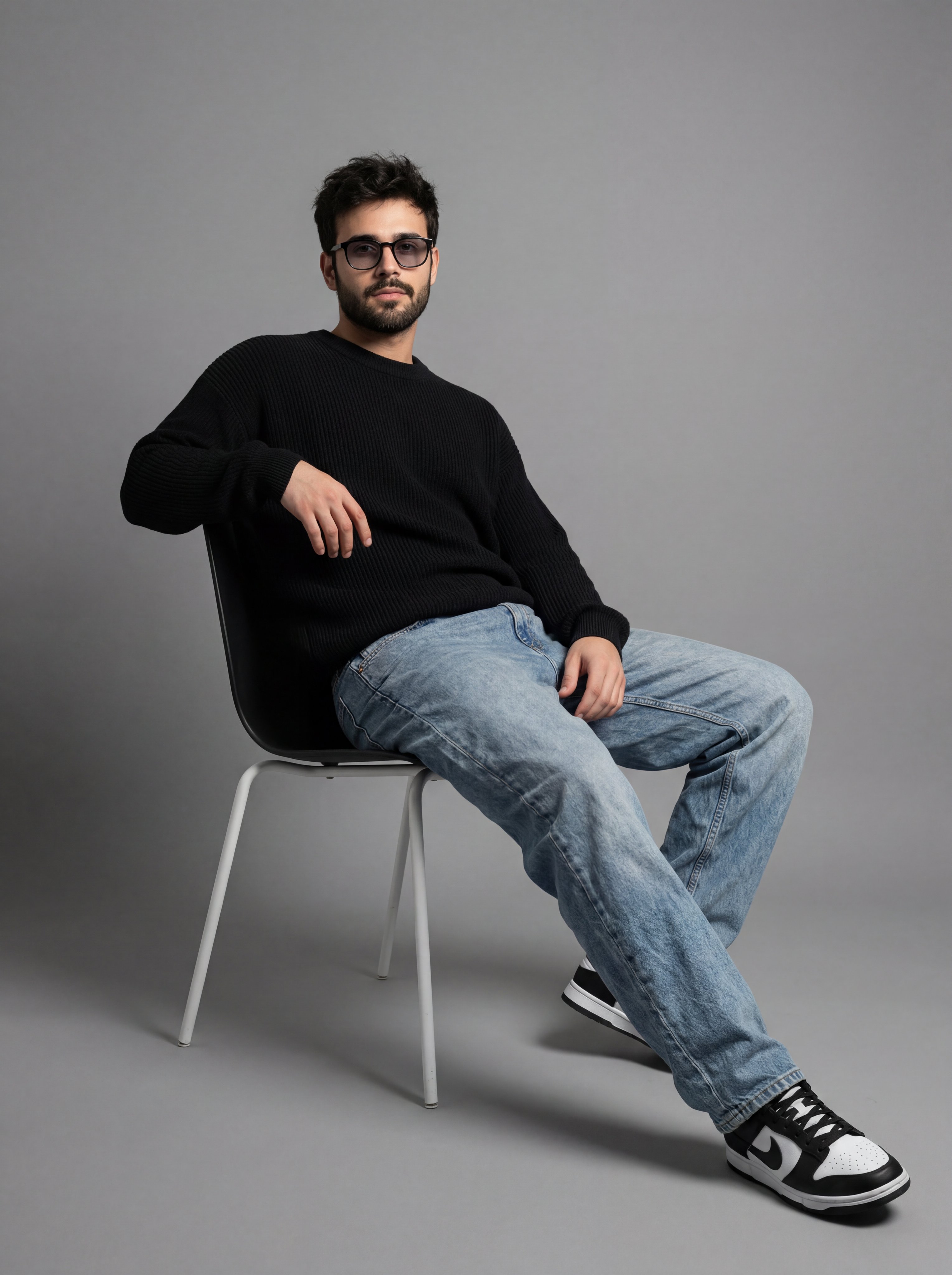 A full-length studio portrait of a young man with dark, messy hair, sit…